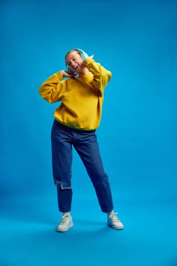 Sarı eşofman altı giyen, kulaklıkla müzik dinleyen ve mavi arka planda dans eden genç bir kızın tam boy görüntüsü. Gençlik kavramı, insani duygular, sıradan moda.