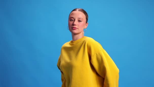 青いスタジオの背景に立って黄色いスウェットシャツを着て ポニーテール ヌードメイクの美しい若い女の子 人間の感情 ライフスタイル カジュアルなファッションの概念 ビデオ — ストック動画