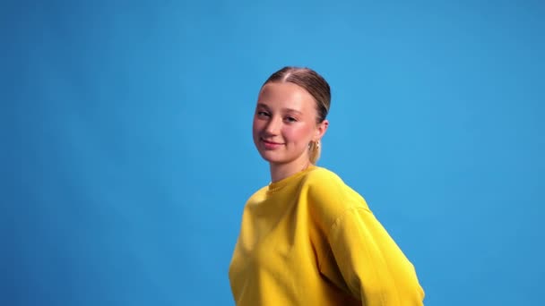 ポニーテール ヌードメイク 黄色のスウェットシャツを着て青いスタジオの背景に微笑む美しい若い女の子 ビデオ 人間の感情 カジュアルなファッション ライフスタイルの概念 — ストック動画