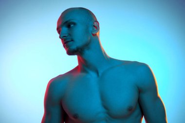 Rahatlatıcı, üstsüz, spor vücutlu yakışıklı genç bir adam, neon ışıkta mavi stüdyo arka planına karşı poz veriyor. Erkek güzelliği, sportif ve sağlıklı yaşam tarzı, vücut bakımı kavramı
