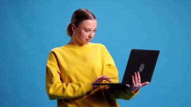 Sarı tişörtlü genç kız dizüstü bilgisayarla çalışıyor, çalışıyor, mavi stüdyo geçmişiyle online alışveriş yapıyor. Gençlik kavramı, insani duygular, sıradan moda. Video, 4k