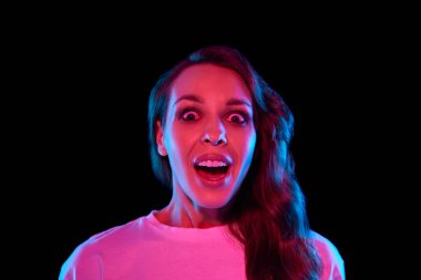 Şok olmuş ve heyecanlı parlak elbiseli genç bir kadın, neon ışıklarıyla siyah stüdyonun arka planında gülümsüyor. Beklenmedik bir sürpriz. Gençlik kavramı, yaşam tarzı, gündelik moda, insani duygular