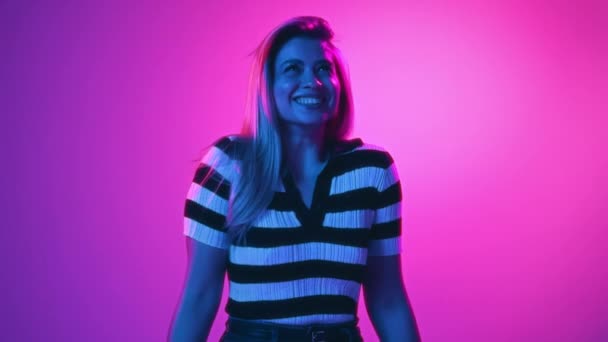 ビデオ 幸せで興奮する若い美しい女性はネオンライトのピンクのスタジオの背景に微笑みます 幸せとは 人間の感情 ファッション 表現の概念 — ストック動画