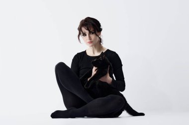 Siyah elbiseli, dumanlı göz makyajlı, beyaz stüdyo arka planında oturan ve sevimli siyah kediyi tutan güzel bir genç kadın. Güzellik kavramı, kadınlık, hayvan teması, moda, sağlık