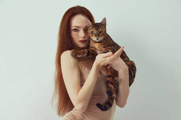 红头发 身穿米色紧身衣的年轻女子的画像 她将孟加拉猫抱在白色的工作室背景之下 开展动物慈善活动 展示人与动物之间的联系 — 图库照片