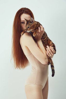 Beyaz stüdyo arka planında Bengal kedisini tutan bej renkli takım elbiseli zarif, güzel, genç bir kızıl kadının portresi. Hayvan teması, kadın güzelliği, hayvan bakımı, arkadaşlık kavramı.