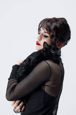 Smoky eyes ve kırmızı dudak makyajı ile güzel genç bir kadın, siyah kıyafetler içinde, beyaz stüdyo arka planında oturan ve sevimli siyah kediyi tutan. Güzellik ve moda kavramı, hayvan bakımı