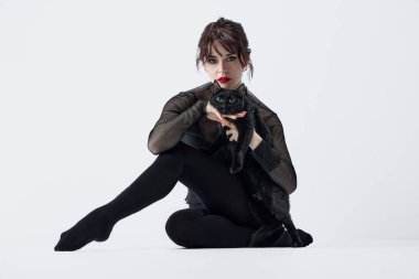 Hayvanlarla bağlantı. Tutkulu, dumanlı gözlü, kırmızı dudaklı, siyah elbiseli, beyaz arka planda siyah kediyle poz veren genç bir kadın. Hayvan teması, hayvan bakımı, güzellik ve moda kavramı