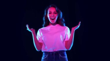 Gülümseyen, neon ışıkta siyah stüdyo geçmişine el çırpan mutlu ve heyecanlı genç bir kadın. - Sürpriz. Mutluluk. Gençlik kavramı, yaşam tarzı, gündelik moda, insani duygular