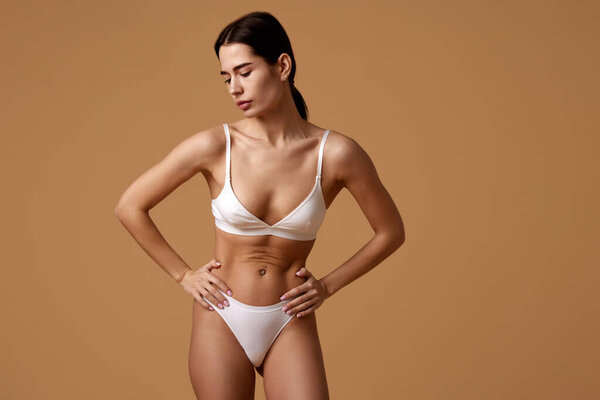 Портрет молодой женщины держит руки на бедрах, подчеркивая ее тонированное, упругое брюхо на фоне песочной цветовой студии. Концепция красоты, спа-процедуры, дерматологические процедуры, косметологический уход.