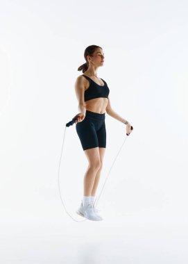 Spor giyim antrenmanında, beyaz stüdyo arka planında izole edilmiş halatla atlama egzersizleri yapan uzun boylu genç bir kadının görüntüsü. Spor, sağlık ve vücut bakımı kavramı, fitness uygulaması