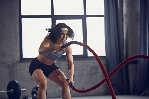 Dynamic portrait of athletic young women in sportswear doing high-intensity rope workouts in motion in gym. Concept of professional sport, bodybuilding, active and healthy lifestyle, energy, action.