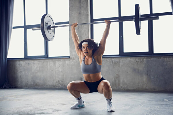 Dynamic portrait of focused young strong woman in sportswear doing squat technique with barbell overhead in motion at gym. Concept of professional sport, active lifestyle, bodybuilding. Ad