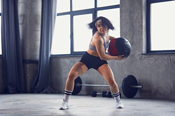 Young athletic curly woman in sportswear passes medicine ball in motion to partner or core strength training in gym. Concept of professional sportsman, healthy lifestyle, workout. Ad