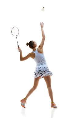 Uzun boylu, sportif bir genç kız, badminton oyuncusu beyaz arka planda izole edilmiş raketle servis yapıyor. Profesyonel spor anlayışı, aktif yaşam tarzı, hobi, oyun