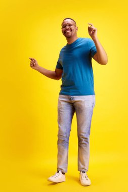 Mutlu, gülümseyen, günlük kıyafetleriyle dans eden, sarı stüdyo geçmişiyle eğlenen Afro-Amerikan bir adamın tam boy görüntüsü. İnsan duyguları kavramı, gündelik moda, yaşam tarzı
