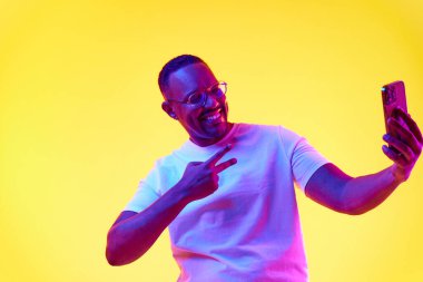 Gözlüklü ve tişörtlü gülümseyen Afro-Amerikan adam, cep telefonuyla selfie çekerken, sarı stüdyo arka planında neon ışığı altında. İnsan duyguları kavramı, gündelik moda, yaşam tarzı