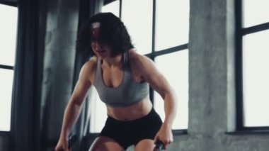 Dinamik videoda genç bir kadın savaş halatı egzersizi yapıyor, endüstriyel jimnastik salonunda dalgalar yaratıyor. Profesyonel spor, vücut geliştirme, aktif ve sağlıklı yaşam tarzı, eğitim. Ad