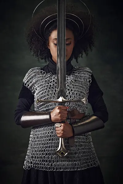 Portrait of young African woman, medieval warrior in chainmail armor with halo-like rings above head holding sword close to face on dark vintage background. Concept of history, comparison of eras