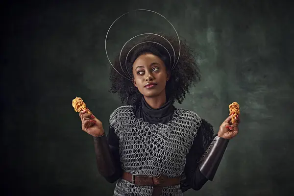 Young African woman, medieval warrior in chainmail armor holding fried chicken against vintage green background. Gourmet food campaign. Concept of comparison of eras, history, creative art, remake