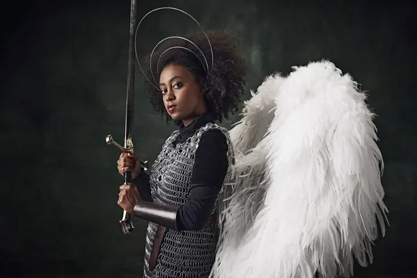 Young African woman, medieval warrior with angel wings, halo above head and chainmail holding sword against vintage dark background. Concept of comparison of eras, history, creative art, remake