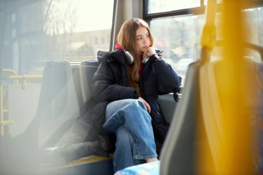 Dreamy looking young woman in headphones sitting in modern tram. Safe transportation. Blurred city streets on background. Concept of public transport, urban lifestyle clipart