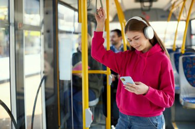 Pembe kapüşonlu genç kız toplu taşıma aracında duruyor, modern tramvay, gülümsüyor, kulaklıkla müzik dinliyor. Güneşli bir gün gezisi. Toplu taşıma kavramı, kentsel yaşam tarzı