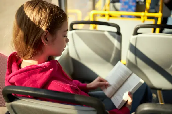 穿着粉色连帽衫的少女坐在公共交通工具 现代有轨电车 公共汽车和阅读书籍中 方便的交通方式 公共交通 城市生活方式的概念 — 图库照片