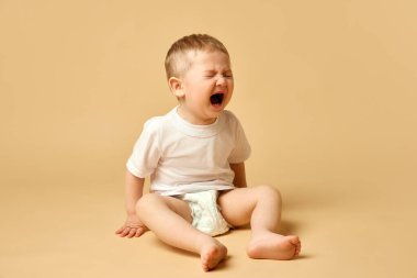 Küçük erkek bebek, tişörtlü çocuk ve bej renkli stüdyo arka planında izole edilmiş bebek bezi bebek bezi oturma ve ağlama. Duygular. Çocukluk ve aile kavramı, bakım, ebeveynlik, bebeklik ve fundalık
