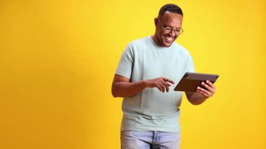 Gözlüklü ve gri tişörtlü gülümseyen Afro-Amerikan adam sarı stüdyo arka planına karşı online alışveriş hizmetleri için tablet kullanıyor. İnsan duyguları kavramı, gündelik moda, yaşam tarzı