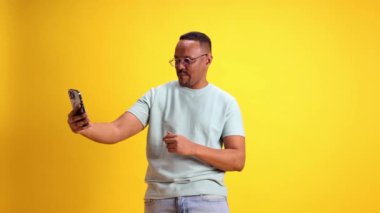 Gözlüklü ve mavi tişörtlü gülümseyen Afro-Amerikan adam sarı stüdyo arka planında cep telefonuyla selfie çekiyor. İnsan duyguları kavramı, gündelik moda, yaşam tarzı