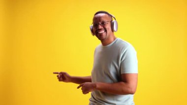 Neşeli, pozitif, rahat, günlük kıyafetler içinde kulaklıkla müzik dinleyen ve sarı stüdyo arka planında dans eden Afro-Amerikan bir adam. İnsan duyguları kavramı, gündelik moda, yaşam tarzı