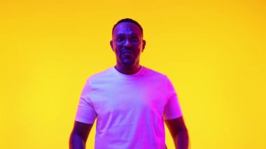 Vay canına. Beyaz tişörtlü etkileyici Afro-Amerikalı adam, neon ışıklı sarı stüdyo arkaplanına karşı şok ve heyecan dolu duygular gösteriyor. İnsan duyguları kavramı, gündelik moda, yaşam tarzı