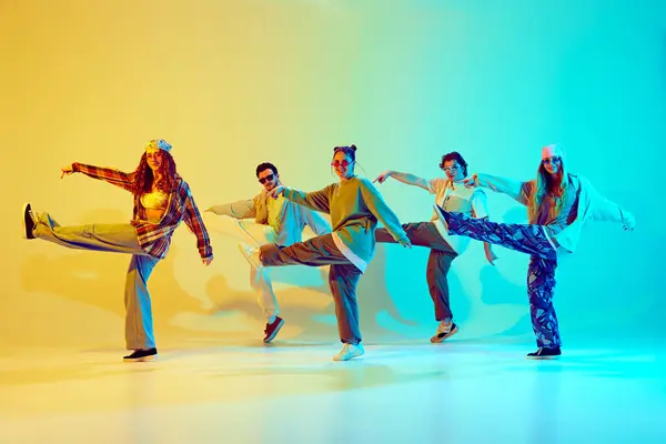 Group of five dancers in casual clothes performing with synchronized poses against gradient green yellow background in neon light. Concept of modern dance style, hobby, active lifestyle, youth culture