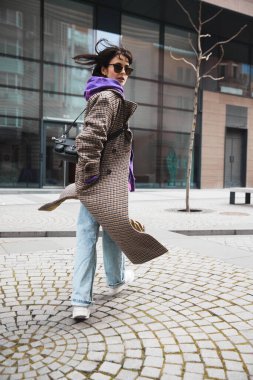 Şık güneş gözlüklü, ceketli ve kapüşonlu şık bir bayan, şehir geçmişine karşı şık bir çanta ile poz veriyor. Rüzgarlı bir hava. Sokak tarzı moda, güzellik, modern trendler