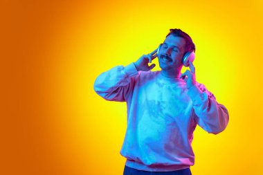 Mutlu, gülümseyen adam kendini rahatlamış hissediyor, kulaklıkla müzik dinliyor, gözleri parlak turuncu arka planda kapalı. İnsan duyguları kavramı, gündelik moda, yaşam tarzı