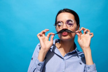 Tişörtlü, takma bıyıklı, gözlüklü, mavi arka planda komik ifadeler kullanan genç bir kadın. Gıda sanatı fotoğrafçılığı, yaratıcılık, acayip bir tarz.