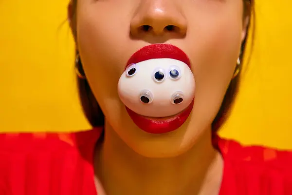 Close-up of womans face with red lipstick, egg on mouth with googly eyes against yellow background. Concept of food pop art photography, creativity, quirky style