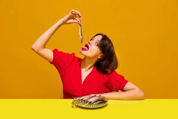 Young woman with smudged lipstick makeup eating fish against yellow background. Expressive dining and food culture. Concept of food pop art photography, creativity, quirky style
