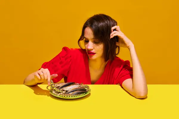 Young woman with smudged lipstick makeup eating fish against yellow background. Impact of food on health and appearance. Concept of food pop art photography, creativity, quirky style