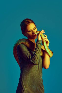 Parlak yeşil elbiseli güzel bir kadın vücudunda yılanla Neon 'daki mavi stüdyo arka planına karşı poz veriyor. Kadın güzelliği kavramı, hayvan teması, özen, zarafet, moda