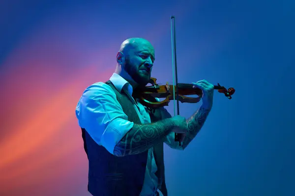 stock image Deep feelings of melody. Young bald man playing violin against blue background in neon with mixed light. Concept of music, talent show, performance, concert, festival, instruments