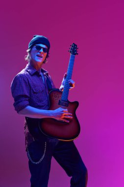 Gündelik kıyafetler ve güneş gözlüğü takan yetenekli, sanatsal genç adam pembe arka planda neon ışıkta gitar çalıyor. Müzik kavramı, yetenek gösterisi, performans, konser, festival, enstrümanlar