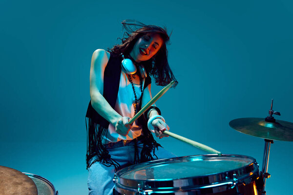 Энергия лопнула. Молодая эмоциональная, активная женщина, музыкант в стильной одежде играет на барабанах на голубом фоне в неоновом свете. Концепция музыки, шоу талантов, перформанс, концерт, фестиваль, инструменты