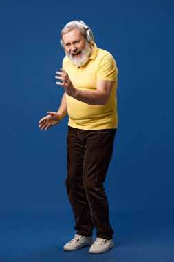 Sarı polo giyen yaşlı sakallı adam kulaklıkla müzik dinliyor ve mavi stüdyo arka planında dans ediyor. Tam boy. İnsan duyguları kavramı, yaşam tarzı, gündelik moda.