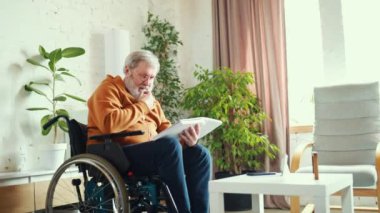 Yaşlı, pozitif bir adam rahat bir oturma odasında, tekerlekli sandalyede oturmuş evde oturup kitap okumaktan zevk alıyor. Sağlık, yaşam tarzı, motivasyon, sağlık, destek kavramı