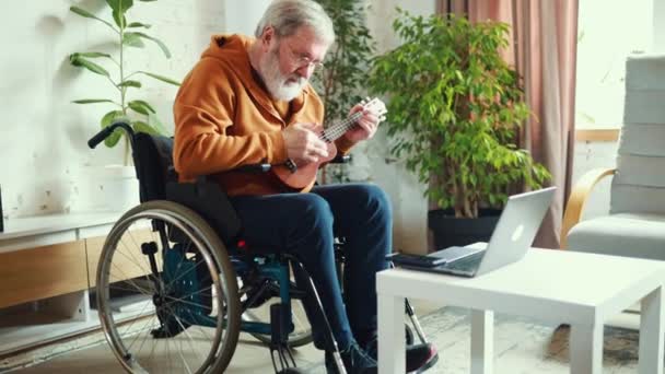 車椅子に座り ラップトップを眺め リビングルームの自宅でウクレレを演奏する老人 音楽の中でリラックスと喜びを見つける ヘルスケア ライフスタイル モチベーション 趣味コンセプト — ストック動画
