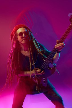 Rastalı, güneş gözlüklü ve bereli bir adamın parlak görüntüsü. Pembe arka planda karışık ışık efektiyle ustaca gitar çalıyor. Müzik kavramı, performans, festival