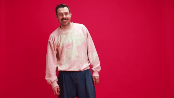 快乐和喜悦 三十多岁的男人穿着粉色运动衫和蓝色牛仔裤 脸上挂着欢乐的笑容 背景是红色的工作室 人类情感 休闲时尚 生活方式 心理概念 — 图库视频影像