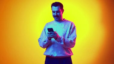 Sweatshirt 'lü ve kotlu gülümseyen genç adam cep telefonuyla neon ışıklı sarı arkaplanda video görüşmesi yapıyor. İnsan duyguları kavramı, gündelik moda, yaşam tarzı